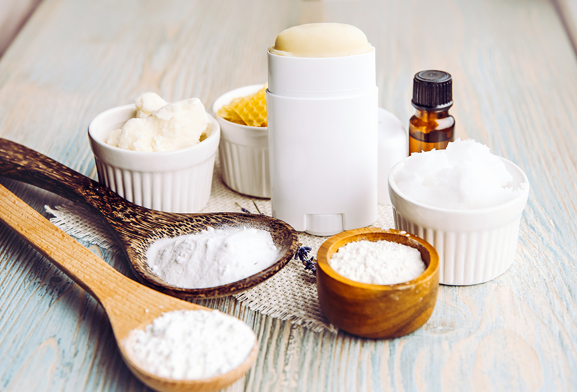 Deodorante solido su un tavolo chiaro in legno, accompagnato da cera d’api in una ciotola e materie prime in polvere su cucchiai di legno.
